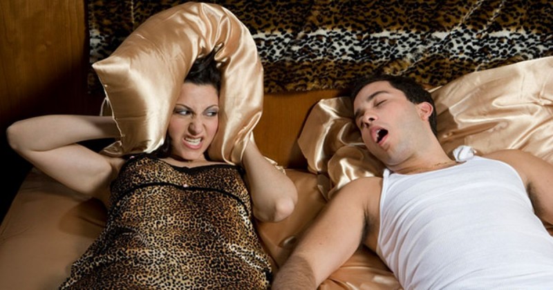 Woman lying on bed next to a snoring man and covering her ears with a pillow